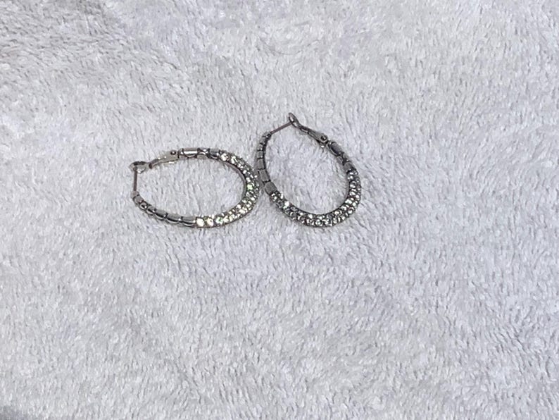 May include: A pair of silver hoop earrings. One earring is in focus, showing a row of small, clear crystals set along the front half of the hoop. The other earring is slightly out of focus, with a similar design. The earrings are on a white, textured surface.