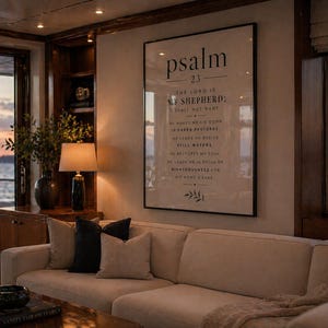 Könnte beinhalten: Ein Wohnzimmer mit einem großen, gerahmten Druck von Psalm 23 über einem beigen Sofa. Der Raum hat einen Holzschrank, eine Lampe und einen Blick auf das Wasser. Dekorative Kissen in Schwarz und Beige liegen auf dem Sofa.