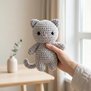 Puede incluir: Una muñeca de gato de ganchillo gris hecha a mano con ojos negros y nariz rosa. El gato tiene orejas pequeñas, cola y brazos extendidos. La muñeca se sostiene en una mano, con un fondo borroso de una mesa y un jarrón.