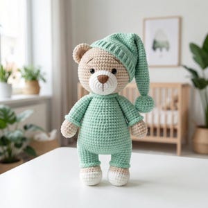 May include: A handmade crochet teddy bear wearing a mint green sleep set and matching nightcap with a pom-pom. The bear has a tan face, arms, and feet, with a white muzzle and a brown nose. The bear is standing on a white surface.