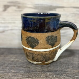 May include: A handmade ceramic mug with a dark blue glazed rim and handle. The body of the mug is tan with black seashell designs and a white stripe. The bottom has a textured, brown pattern.
