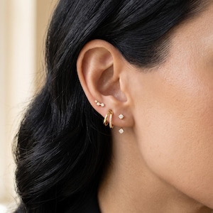 Op de afbeelding: Close-up van een oor versierd met meerdere gouden oorbellen. Het oor heeft een kleine cluster van drie studs, een paar diamanten studs en gouden oorringen met kleine, sprankelende steentjes.