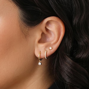 May include: A person's ear is shown with three gold earrings. There's a small stud, a small hoop with crystals, and a larger hoop with a dangling gem. The earrings are set against a dark background. The gems are teardrop-shaped.