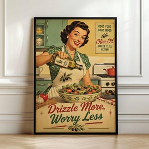 Op de afbeelding: Een ingelijste kunstdruk met een glimlachend persoon in een groene jurk met stippen en een wit schort die olijfolie over een salade giet. De tekst luidt: Good Food Good Mood Olive Oil Makes It All Better! en Drizzle More, Worry Less.