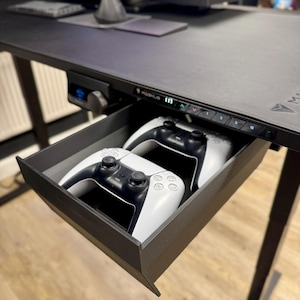 May include: A black under-desk storage drawer attached to a desk, containing two white and black wireless gaming controllers. The desk features a control panel with the text MAGNUS and numbered buttons for height adjustment settings.