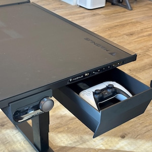 Op de afbeelding: Een zwart sta-bureau met een mat oppervlak en het woord MAGNUS op de rand. Het bureau heeft een bedieningspaneel met een ronde draaiknop en een uitschuifbare lade met daarin een wit-zwarte draadloze gamecontroller.
