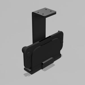 May include: A matte black gaming console mount featuring a vertical bracket with four screw holes at the top and a horizontal base designed to hold a handheld gaming device securely in place. The structure is solid, angular, and uniform in colour.