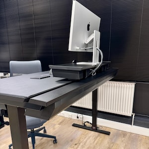 May include: A silver all-in-one computer with a black Apple logo on the back, positioned on a black monitor stand atop a black standing desk. The desk features a black leather-like mat, and a gray office chair is tucked underneath the desk.