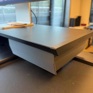 Op de afbeelding: Een zwarte monitorstandaard met een getextureerd bovenoppervlak en een grijze basis. De standaard heeft een slanke, schuine voorrand en staat op een bureau, waarbij het een computermonitor ondersteunt die bovenaan het beeld zichtbaar is.