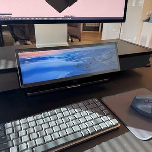 Peut inclure: Un moniteur secondaire large et rectangulaire affichant un paysage de montagne enneigé, placé sous un écran d'ordinateur plus grand. Un clavier mécanique gris avec des lettres blanches et une souris d'ordinateur gris foncé sur un tapis sont au premier plan.