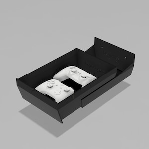Könnte beinhalten: Eine mattschwarze, rechteckige Ablageschale mit einer erhöhten Rückwand, die zwei kleine kreisförmige Löcher aufweist. In der Schale liegen zwei weiße, kabellose Videospiel-Controller nebeneinander und zeigen ihre Tasten und Analogsticks.