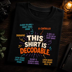 Op de afbeelding: Een zwart t-shirt met de tekst THIS SHIRT IS DECODABLE in grote crème en oranje letters. Rondom de tekst staan kleurrijke pijlen en labels, waaronder CLOSED SYLLABLE, R-CONTROLLED, DIGRAPHS en andere taalkundige aantekeningen.