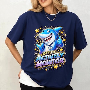 Könnte beinhalten: Marineblaues T-Shirt mit einem Cartoon-Hai-Design. Der Hai ist blau und weiß, gibt einen Daumen hoch und ist von gelben Sternen umgeben. Der Text "OFF TO ACTIVELY MONITOR" ist in einer bunten Schriftart geschrieben.