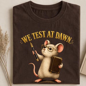 Może przedstawiać: Ciemnobrązowa koszulka z myszą z kreskówki w okularach, trzymającą pióro i książkę. Tekst "WE TEST AT DAWN" jest wydrukowany na złoto nad myszą. Koszulka jest wykonana z miękkiego materiału.