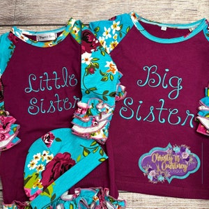 May include: Two long-sleeved shirts with floral print on the sleeves. The shirts are burgundy with white and blue floral print on the sleeves. The left shirt says "Little Sister" in blue embroidery. The right shirt says "Big Sister" in blue embroidery. A matching floral print hat is included.