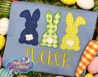 May include: A light blue t-shirt with three embroidered bunny appliques in blue, green, and yellow. The bunnies have a gingham pattern and the text "Tucker" is embroidered in yellow below them.