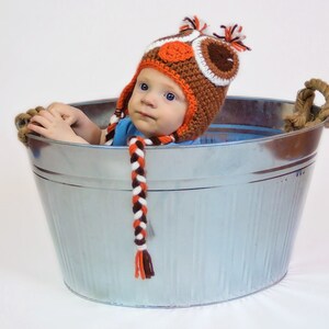 K&ouml;nnte beinhalten: Ein Baby tr&auml;gt eine braune, orange und wei&szlig;e, geh&auml;kelte M&uuml;tze mit Eulenmotiv und sitzt in einer silbernen Metallwanne mit Seilgriffen.