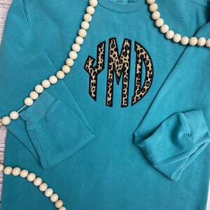 May include: Teal sweatshirt with a black and brown leopard print monogram "YMD" on the front. The sweatshirt is laying flat on a wooden surface with a string of wooden beads.