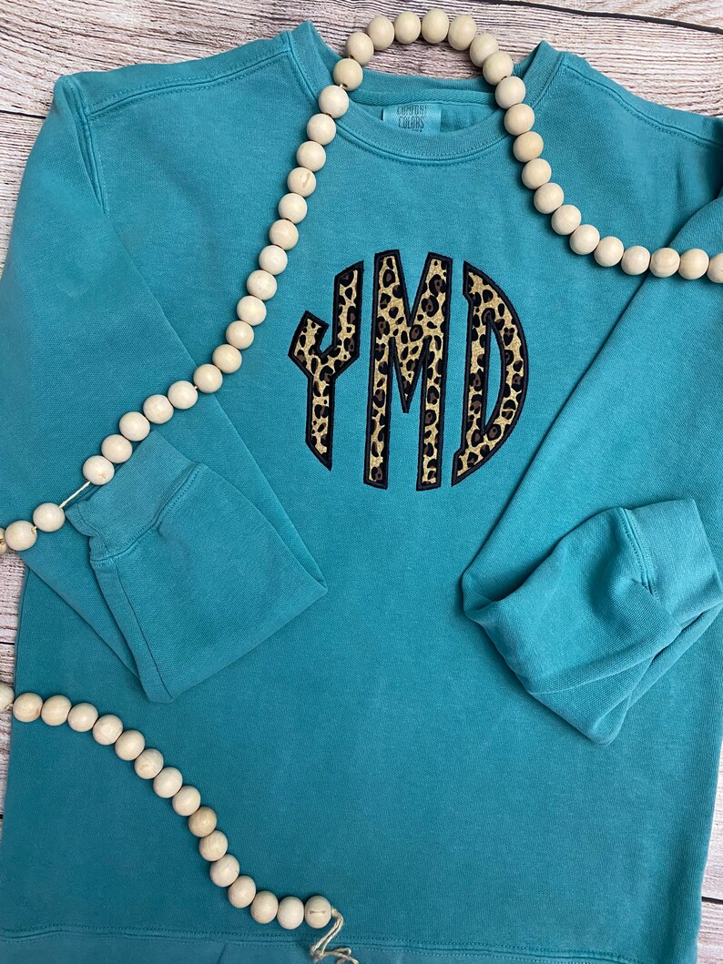 May include: Teal sweatshirt with a black and brown leopard print monogram "YMD" on the front. The sweatshirt is laying flat on a wooden surface with a string of wooden beads.