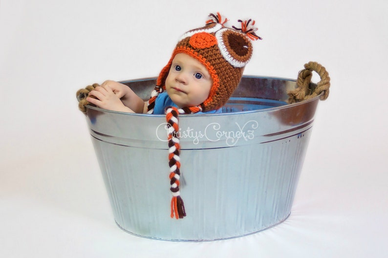 K&ouml;nnte beinhalten: Ein Baby tr&auml;gt eine orangefarbene, braune und wei&szlig;e geh&auml;kelte Eulenm&uuml;tze und sitzt in einer silbernen verzinkten Wanne mit Seilgriffen. Die M&uuml;tze hat einen langen geflochtenen Schwanz.