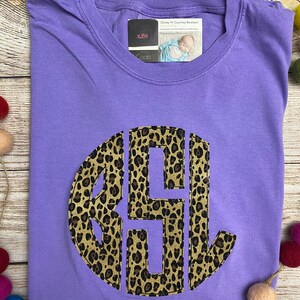 May include: Purple t-shirt with a large, round, leopard print monogram. The monogram is in the center of the shirt and is a dark brown color. The shirt is on a wooden surface with colorful balls scattered around it.