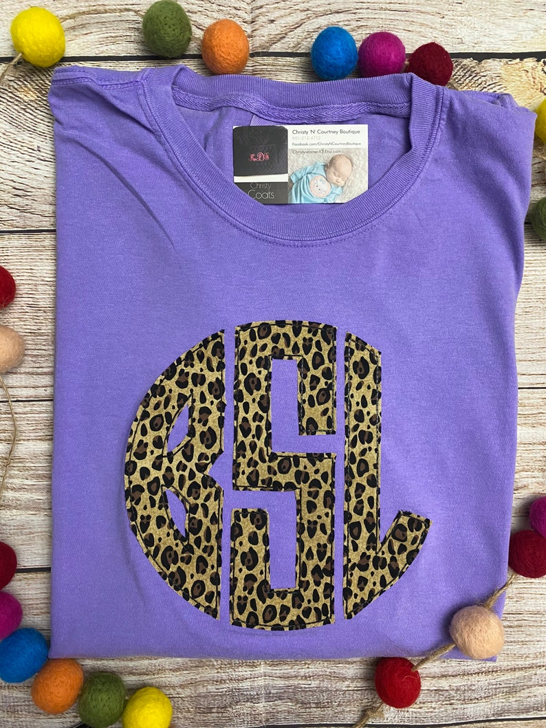 May include: Purple t-shirt with a large, round, leopard print monogram. The monogram is in the center of the shirt and is a dark brown color. The shirt is on a wooden surface with colorful balls scattered around it.
