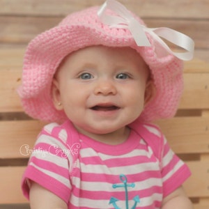 May include: A baby girl wearing a pink and white striped shirt with an anchor design and the name "Mckenna" embroidered on it. She is also wearing a pink crocheted sun hat with a white ribbon.