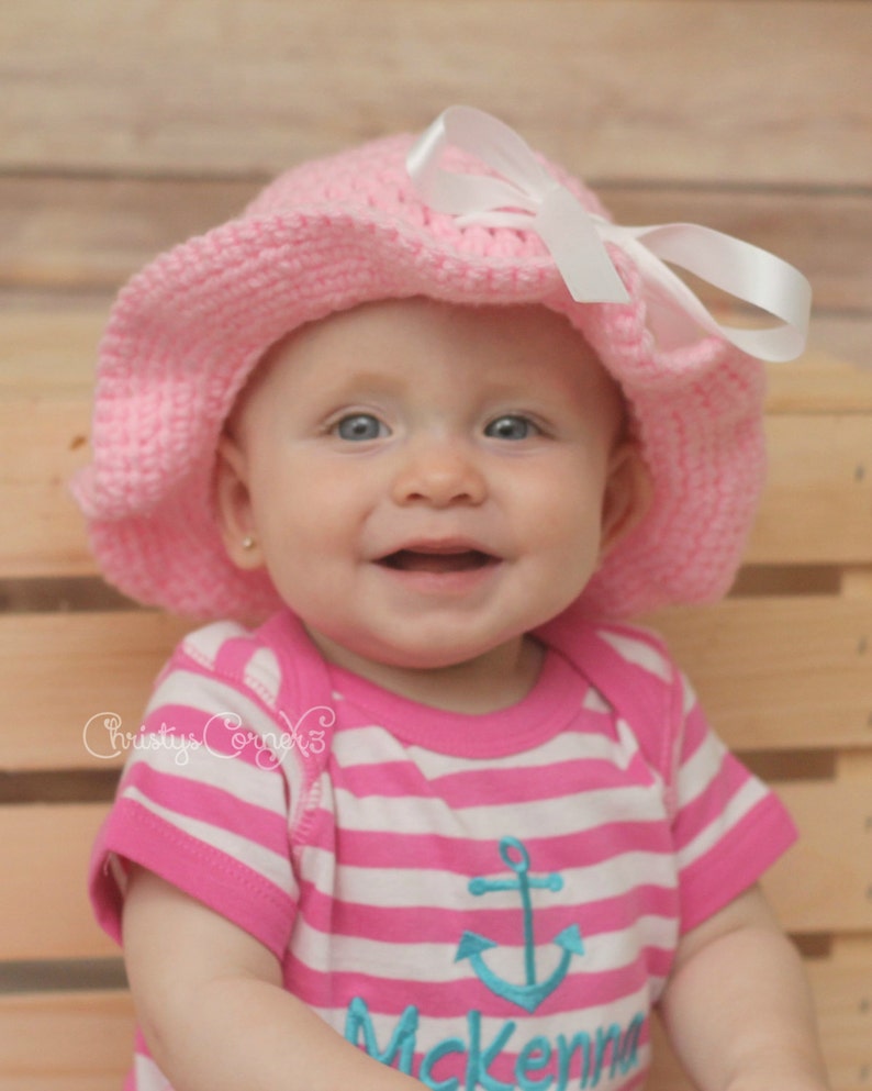 May include: A baby girl wearing a pink and white striped shirt with an anchor design and the name "Mckenna" embroidered on it. She is also wearing a pink crocheted sun hat with a white ribbon.