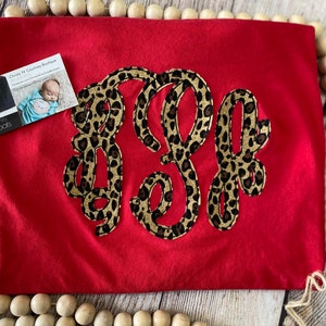 May include: Red t-shirt with a large monogram in a leopard print design. The monogram is made up of the letters "K" and "E".