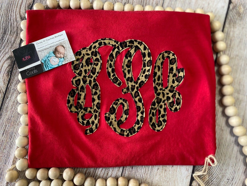 May include: Red t-shirt with a large monogram in a leopard print design. The monogram is made up of the letters "K" and "E".