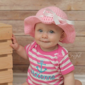 May include: A baby girl wearing a pink crocheted sun hat with a white ribbon and a pink and white striped shirt with an anchor and the name "Mckenna" embroidered on it.