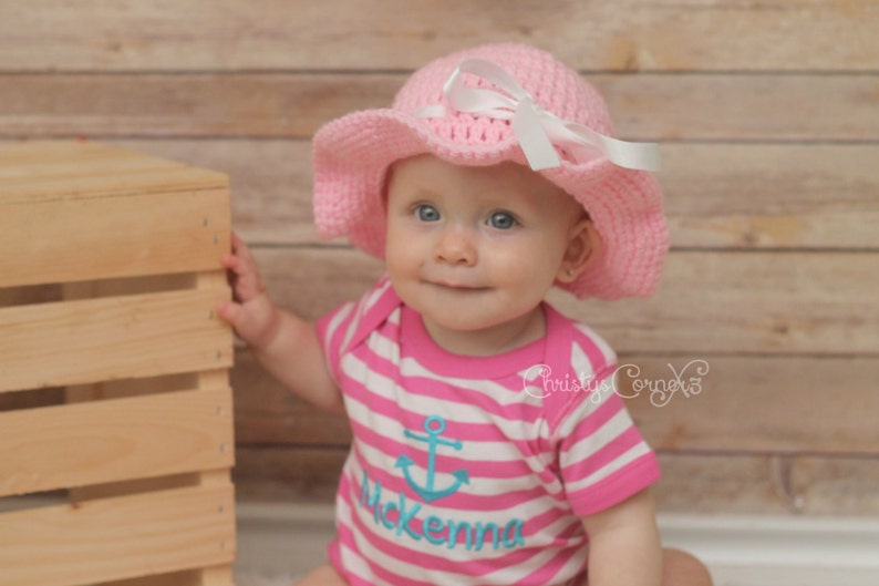 May include: A baby girl wearing a pink crocheted sun hat with a white ribbon and a pink and white striped shirt with an anchor and the name "Mckenna" embroidered on it.