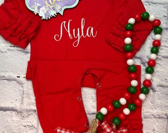 May include: Red long-sleeve baby onesie with white ruffle trim and the name "Myla" embroidered on the front. A red, white, and green beaded garland with a red and white checkered bow and a wooden tag that says "Merry" is included.