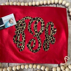 May include: Red t-shirt with a large monogram in a leopard print design. The monogram is in a cursive font and is centered on the shirt. The shirt is lying flat on a wooden surface.