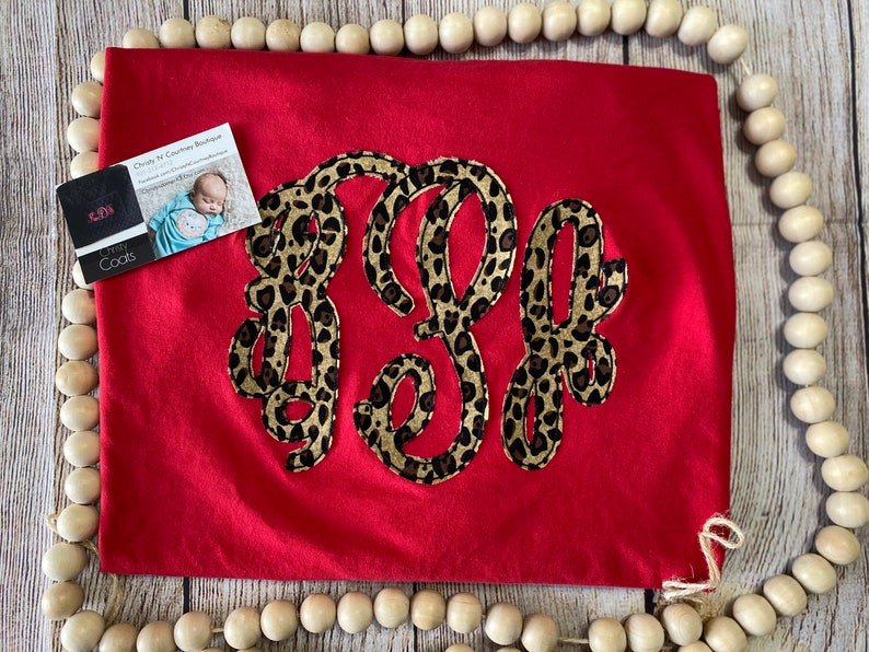 May include: Red t-shirt with a large monogram in a leopard print design. The monogram is in a cursive font and is centered on the shirt. The shirt is lying flat on a wooden surface.