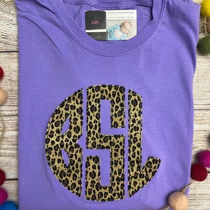 May include: Purple t-shirt with a large, round, leopard print monogram. The monogram is in brown and black. The shirt is on a wooden surface with colorful balls.