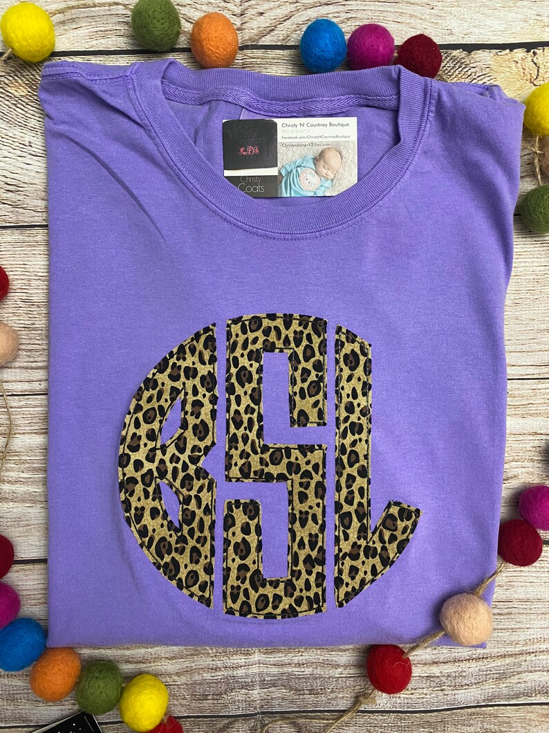 May include: Purple t-shirt with a large, round, leopard print monogram. The monogram is in brown and black. The shirt is on a wooden surface with colorful balls.