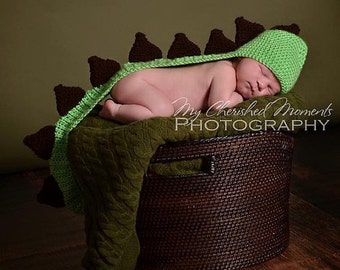 May include: A newborn baby wearing a green and brown crocheted dinosaur costume is sleeping in a brown wicker basket. The baby is wearing a green crocheted hat with brown spikes. The basket is sitting on a wooden floor.