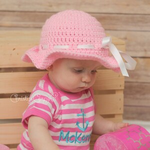 May include: A pink crocheted sun hat with a white ribbon and a white bow. The hat is worn by a child wearing a pink and white striped shirt with a blue anchor and the name "Mcken" embroidered on it.