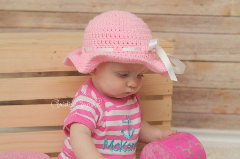 May include: A pink crocheted sun hat with a white ribbon and a white bow. The hat is worn by a child wearing a pink and white striped shirt with a blue anchor and the name "Mcken" embroidered on it.