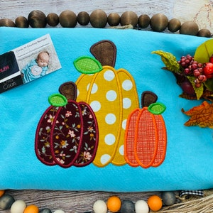 May include: A blue shirt with three embroidered pumpkins. The pumpkins are different colors and patterns. The center pumpkin is yellow with white polka dots. The pumpkin on the left is brown with a floral pattern. The pumpkin on the right is orange with a checkered pattern.