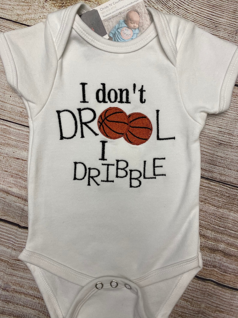 May include: White baby onesie with the embroidered text "I don't DROOL I DRIBBLE" in black. Two brown basketballs are embroidered between the words. The onesie has short sleeves and snaps at the bottom.