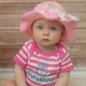May include: A baby girl wearing a pink crocheted sun hat with a white bow and a pink and white striped shirt with a blue anchor and the name "Mckenna" embroidered on it.