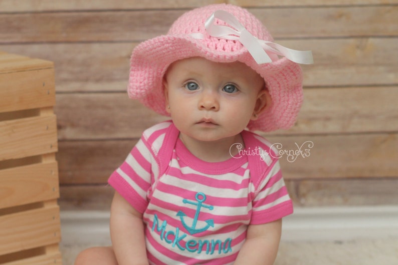 May include: A baby girl wearing a pink crocheted sun hat with a white bow and a pink and white striped shirt with a blue anchor and the name "Mckenna" embroidered on it.