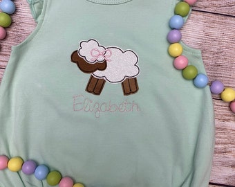 May include: A light green baby romper with a sheep applique. The sheep has a white body, brown legs, and a chocolate-colored head with a pink bow. The name "Elizabeth" is embroidered below the sheep. Ruffled sleeves and trim.