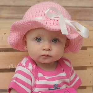 May include: A baby girl wearing a pink crocheted sun hat with a white ribbon and a pink and white striped shirt.