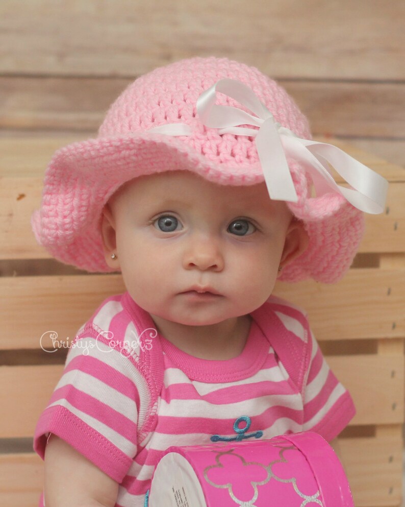 May include: A baby girl wearing a pink crocheted sun hat with a white ribbon and a pink and white striped shirt.