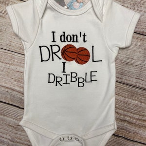 May include: White baby onesie with the text "I don't drool I dribble" in black, with two embroidered basketballs. The onesie has short sleeves and snap closures at the bottom.