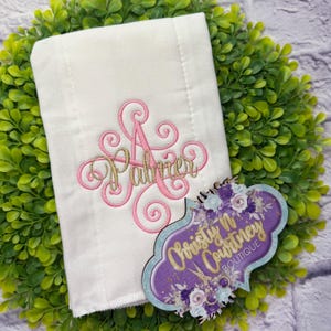 May include: A white cloth with a pink and gold embroidered design. The design includes the letter "A" and the name "Palmer" in a cursive font. A decorative tag with the text "Christy N Courtney Boutique" is also present.