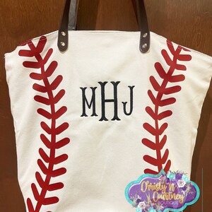May include: Cream-colored tote bag with red baseball stitching design and brown leather handles. Features the embroidered initials "MHJ" in black. The bag has a decorative tag with the text "Christy n Courtney Boutique".